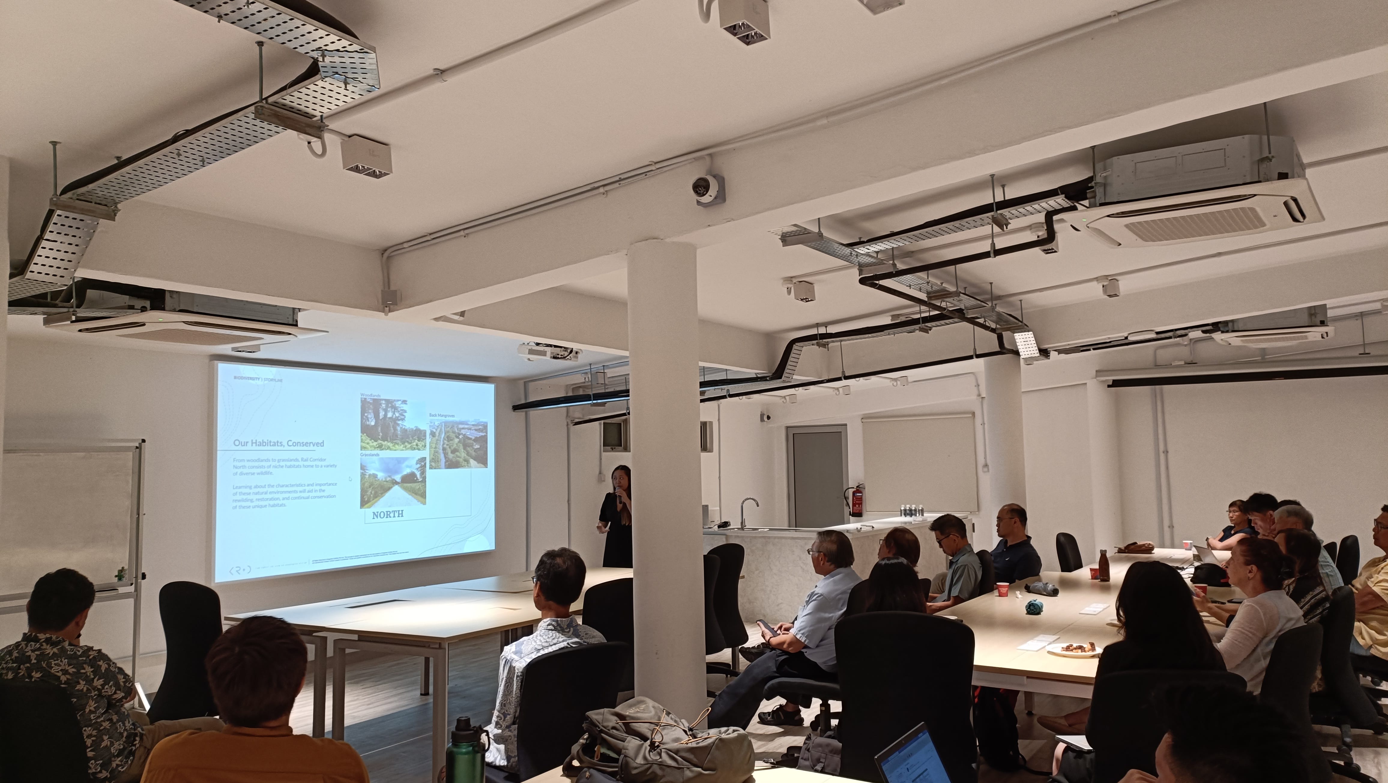People seated in a meeting room watching a presentation projected on a screen reading "Our Habitats, Conserved".